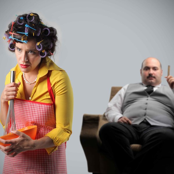 Frau mit Kochschürze und Locken im Haar mit Mann der auf Stuhl sitzt